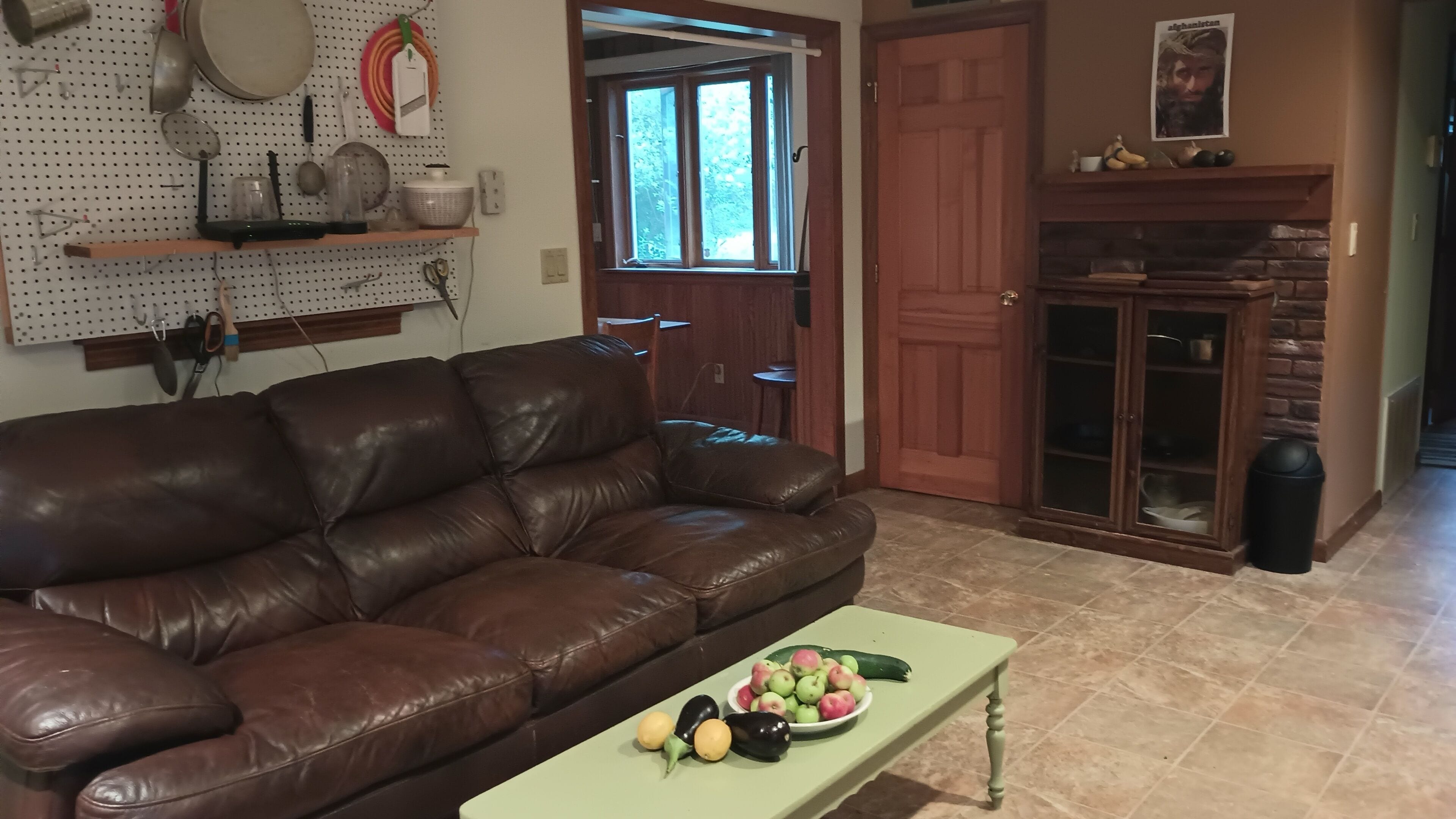 A living room with a brown leather couch, a green coffee table with fruits, a pegboard with kitchen utensils, and a fireplace.