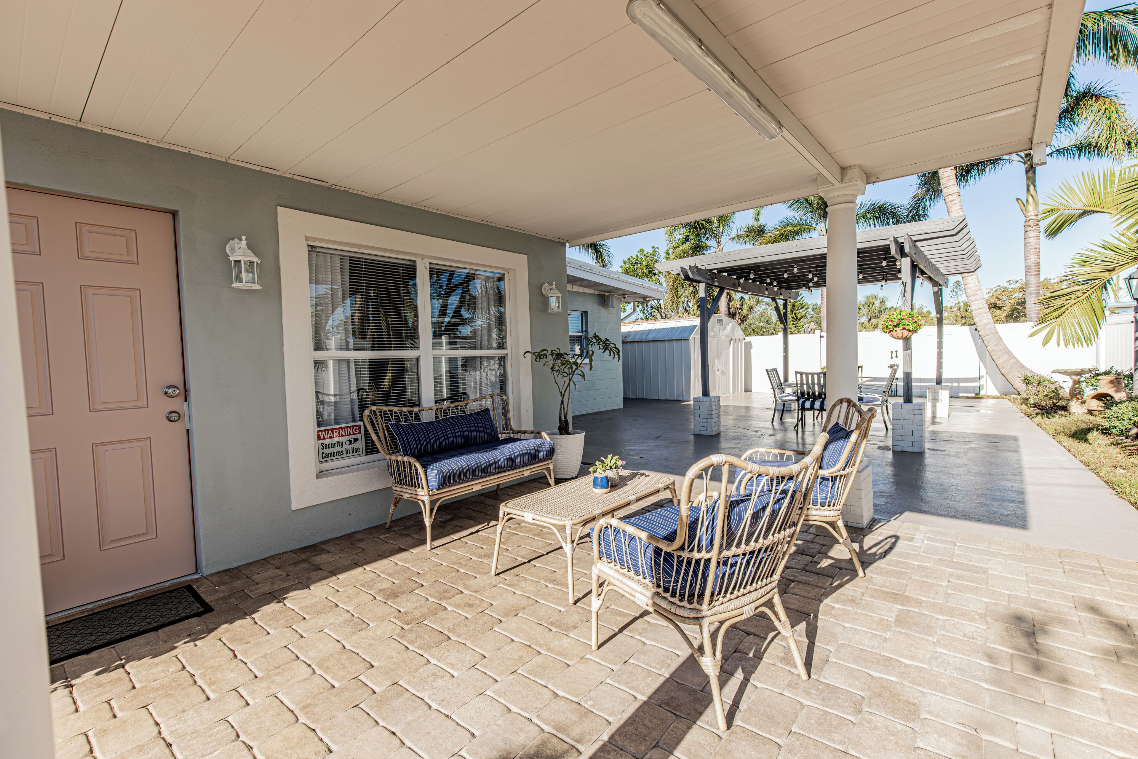A covered patio area with wicker furniture, a security warning sign, and a view of palm trees.