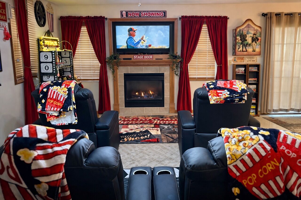 A living room with a fireplace, two black leather chairs, a television mounted above the fireplace, and red curtains.