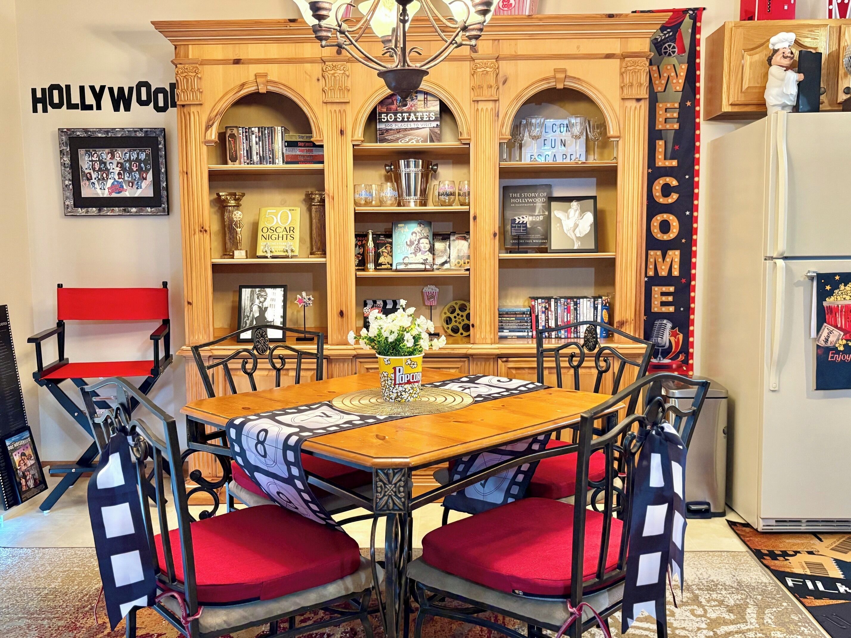 A dining area with a wooden table, red cushioned chairs, a popcorn stand, and a bookshelf with various books and decorations.