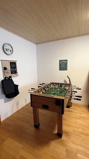 A room with a foosball table, a clock, a Jack Daniel's sign, and a black backpack on the wall.