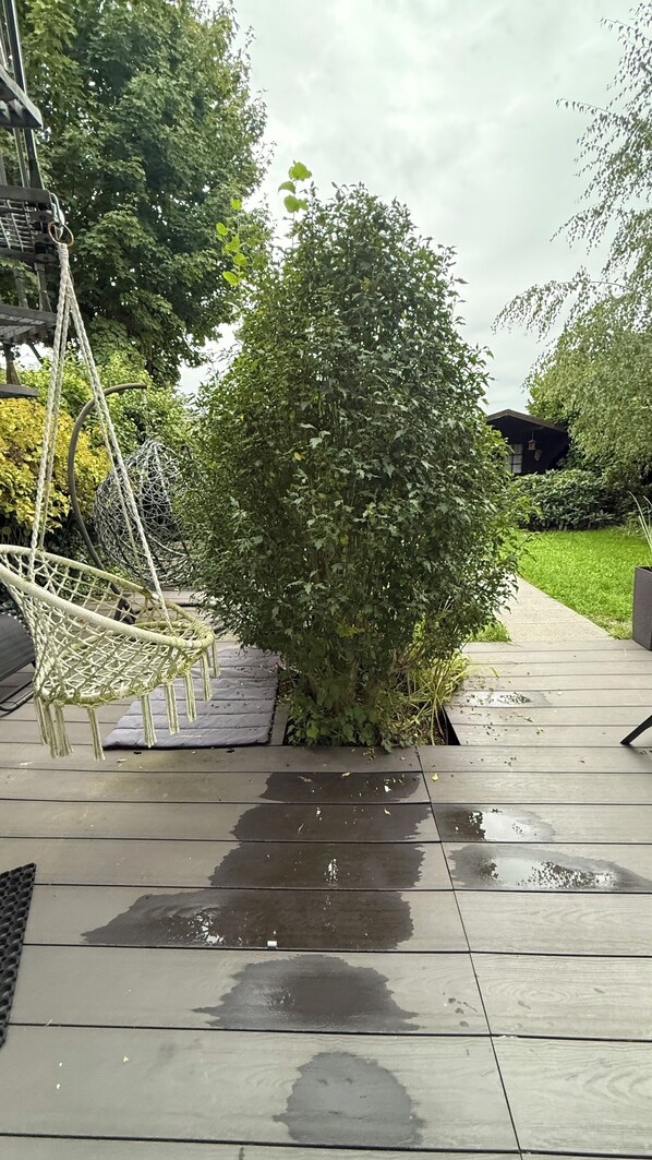 A wooden deck with a small bush, a swing, and a pathway leading to a house.
