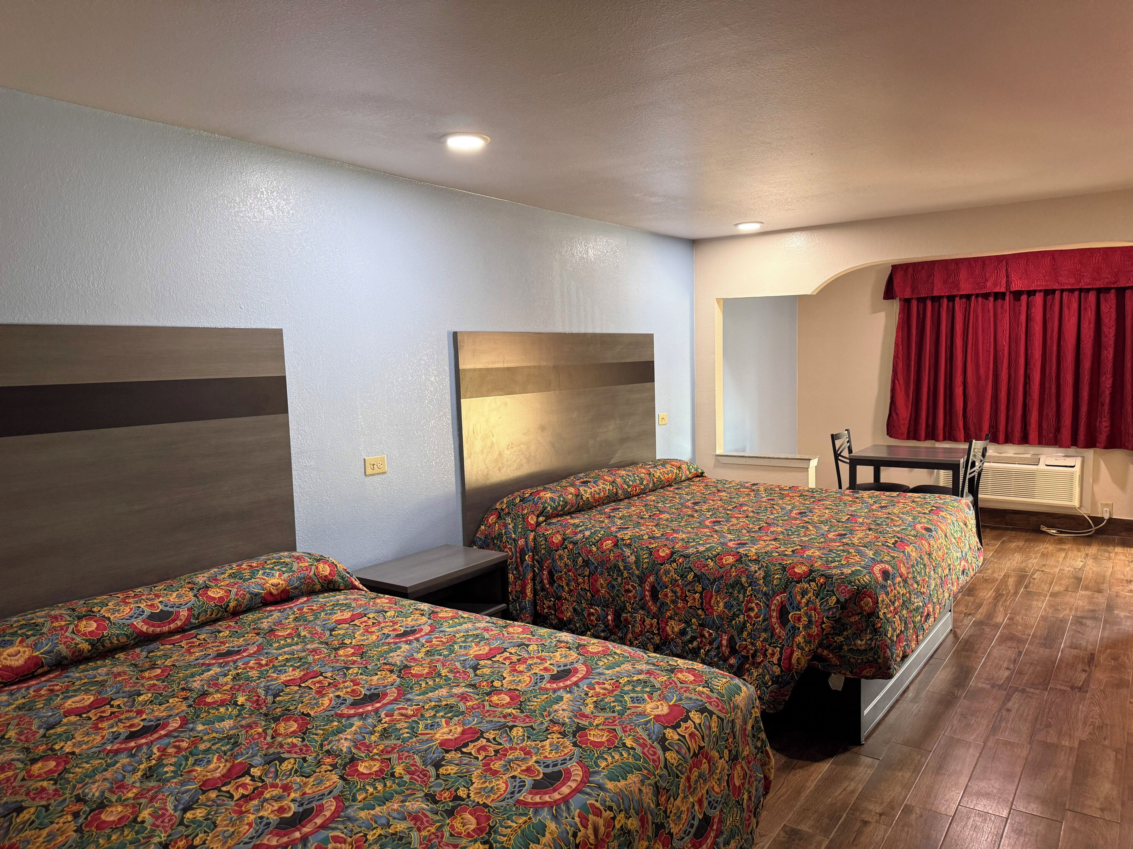 Ein Hotelzimmer mit zwei Betten, einem kleinen Tisch, einem Stuhl und einem Fenster mit roten Vorhängen.