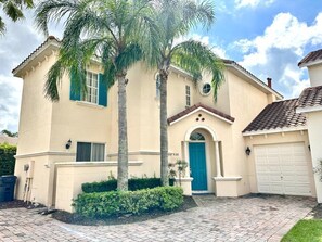 A two-story house with a blue door, a garage, and a palm tree in the front yard. - Tuscan Hills 5BR Villa | Private Pool & Spa (Davenport)