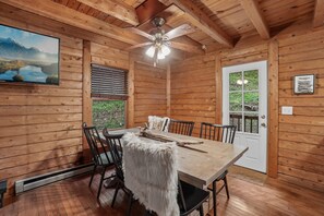 A wooden cabin interior with a dining table, chairs, a ceiling fan, and a large window with blinds. - Wolf Laurel Cabin with Hot Tub Near Skiing (Mars Hill)