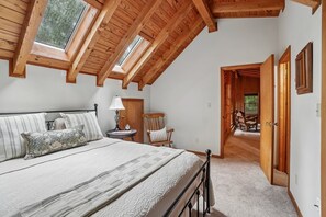 A bedroom with a sloped ceiling, a bed with a quilt, a nightstand with a lamp, a chair, and a view through an open door to another room. - Wolf Laurel Cabin with Hot Tub Near Skiing (Mars Hill)