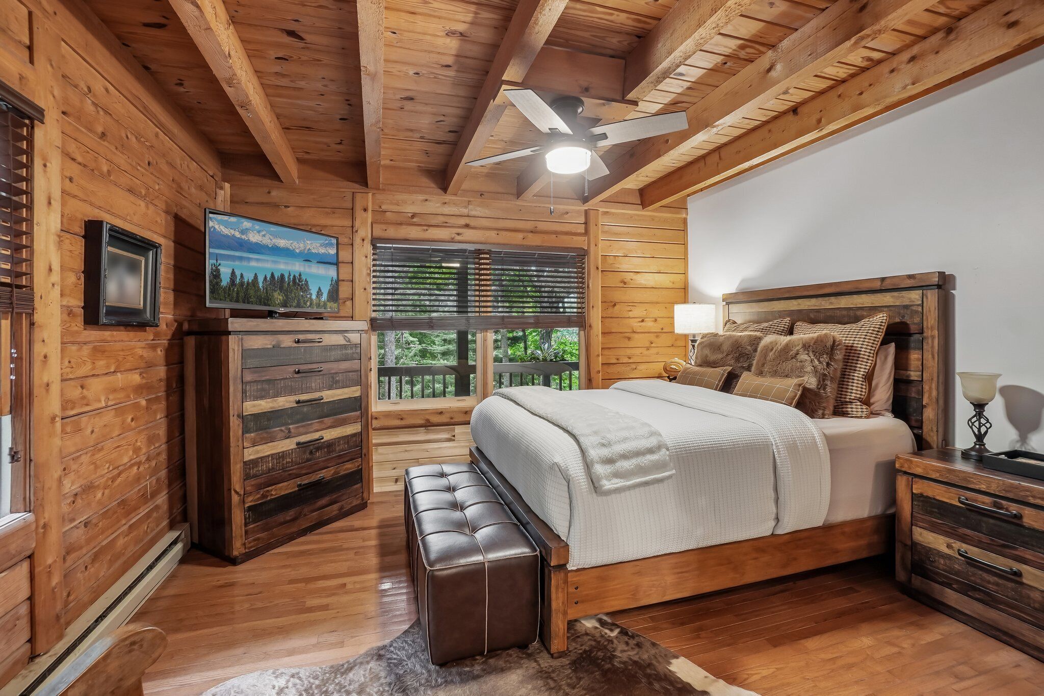 A wooden cabin bedroom with a bed, a bench, a television, and a window with blinds.
