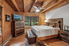 A wooden cabin bedroom with a bed, a bench, a television, and a window with blinds. - Wolf Laurel Cabin with Hot Tub Near Skiing (Mars Hill)
