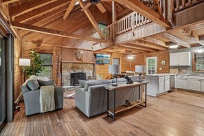 A spacious living area with a stone fireplace, grey sofas, a wooden dining table, and a kitchen with white cabinets. - Wolf Laurel Cabin with Hot Tub Near Skiing (Mars Hill)