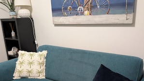Un sofá azul aguamarina con un cojín estampado con hojas de palma y un cojín oscuro, una lámpara blanca, una pequeña estantería con objetos decorativos y un cuadro de una bicicleta en la playa.