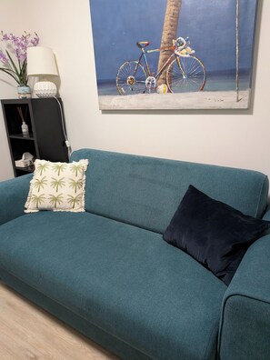 Ein blaugrünes Sofa mit einem Kissen mit Palmenmuster und einem dunklen Kissen, eine weiße Lampe, ein kleines Regal mit Dekoration und ein Bild von einem Fahrrad am Strand.