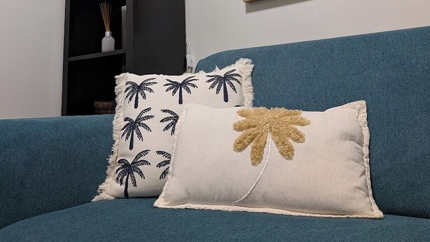 Un sofá azul turquesa con dos cojines decorativos, un cuadro abstracto colgado en la pared, una lámpara blanca y una planta pequeña sobre una mesita de noche negra.