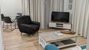 Una sala moderna con un televisor de pantalla plana, un sillón negro, un sofá azul, una mesa de centro de madera y una zona de comedor con una mesa y sillas de vidrio.