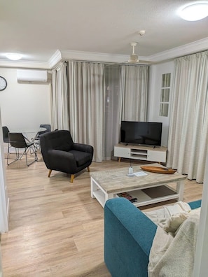 Ein modernes Wohnzimmer mit Flachbildfernseher, einem schwarzen Sessel, einem blauen Sofa, einem Holztisch und einem Essbereich mit Glastisch und Stühlen.