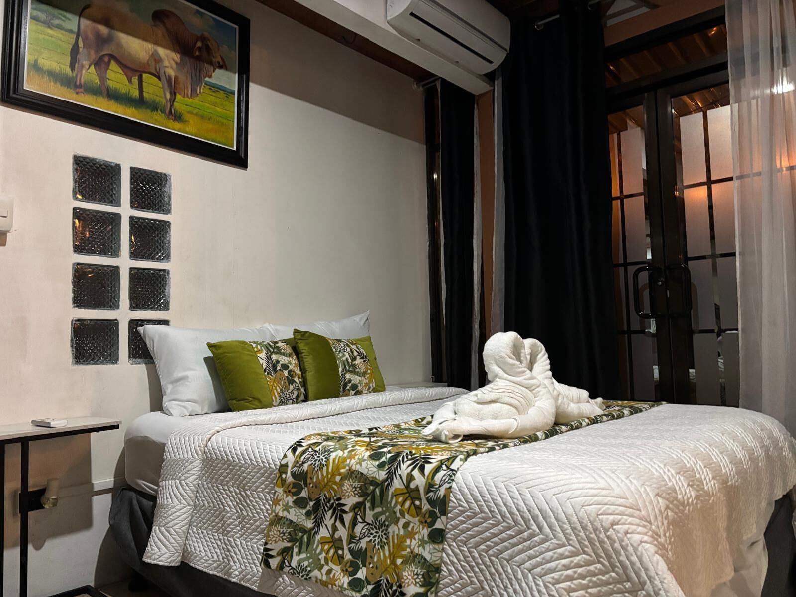 A hotel room with a bed, a painting, a window with blinds, and a bedside table.