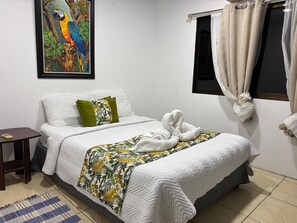 A neatly made bed with a white quilt, a green pillow, and a tropical leaf-patterned throw. A framed colorful parrot painting adorns the wall above the bed.