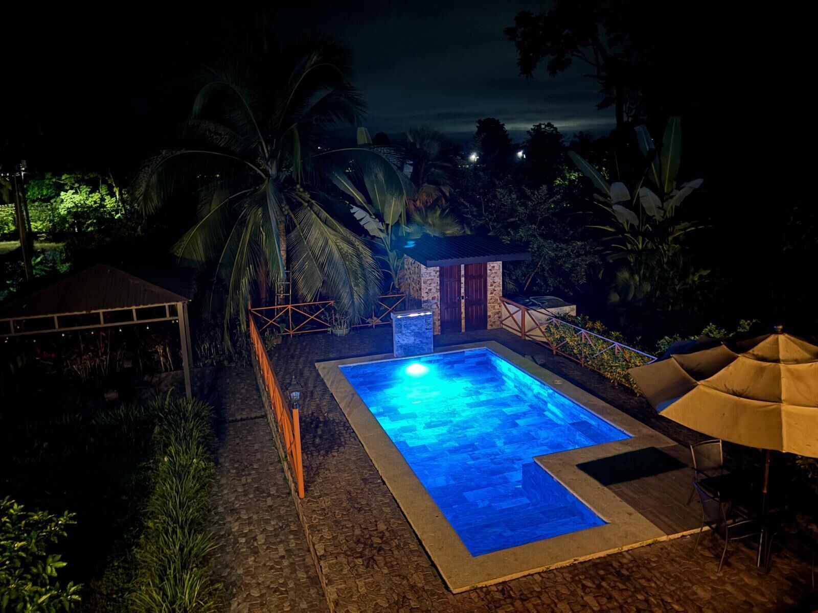 Uma piscina noturna com borda de pedra, rodeada por palmeiras e um pequeno edifício.