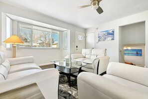 A modern living room with a glass coffee table, white leather sofas, a ceiling fan, and a window with blinds.