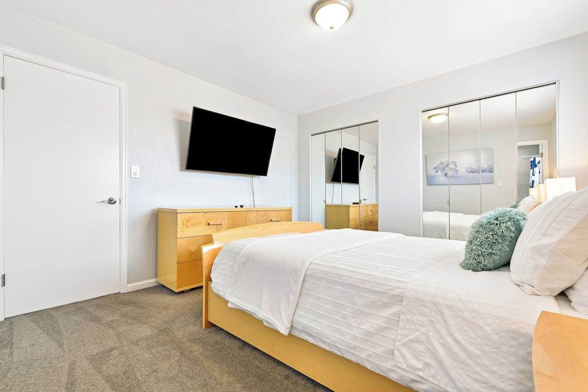 Ein Schlafzimmer mit einem Bett, einer Holzkommode, einem an der Wand befestigten Fernseher und einem großen Spiegel.