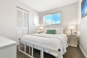 Un dormitorio con cama, mesitas de noche, una ventana y una puerta.