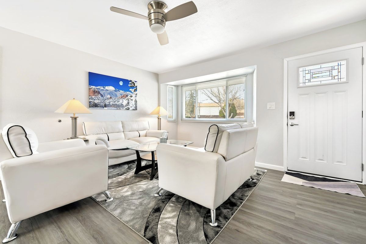 A modern living room with a white sofa set, a coffee table, a ceiling fan, a large window, and a door with a geometric design.