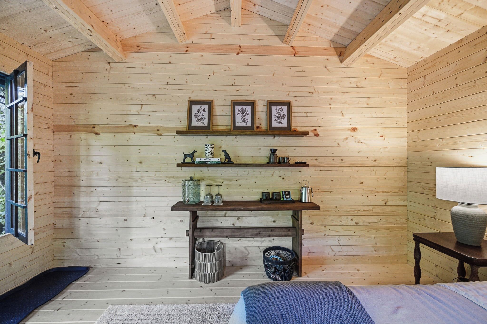 Een houten blokhut van binnen met een bed, een plank met decoratieve objecten, en een raam.