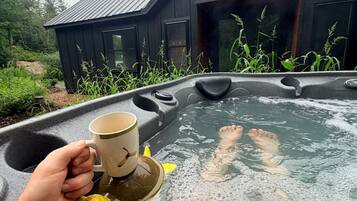 A hot tub with a person holding a mug, a yellow duck toy, and a wooden building in the background.