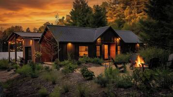 A cabin with a fire pit, surrounded by dense forest, at dusk.