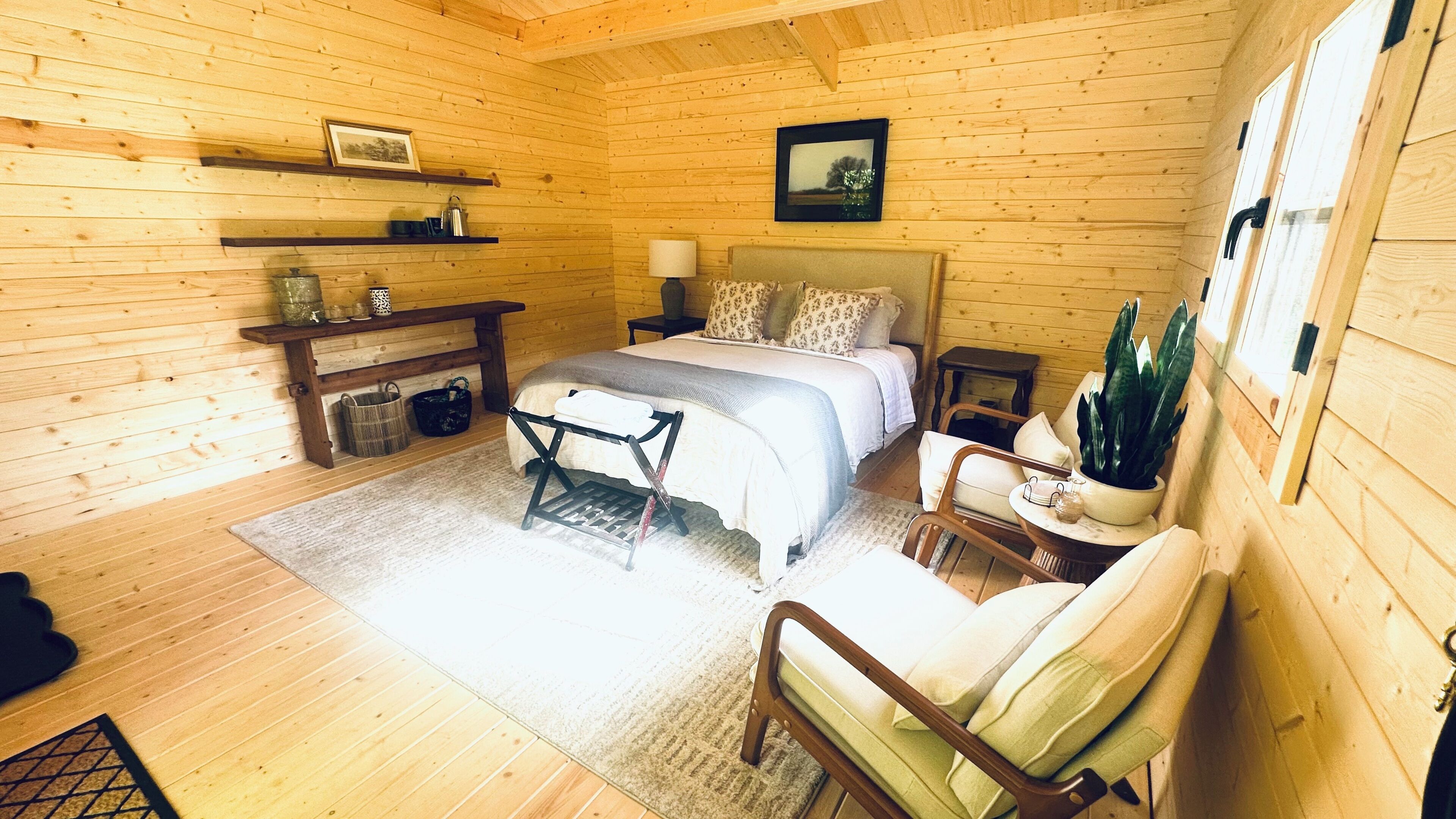 A wooden cabin bedroom with a bed, bedside table, lamp, chair, and a potted plant.