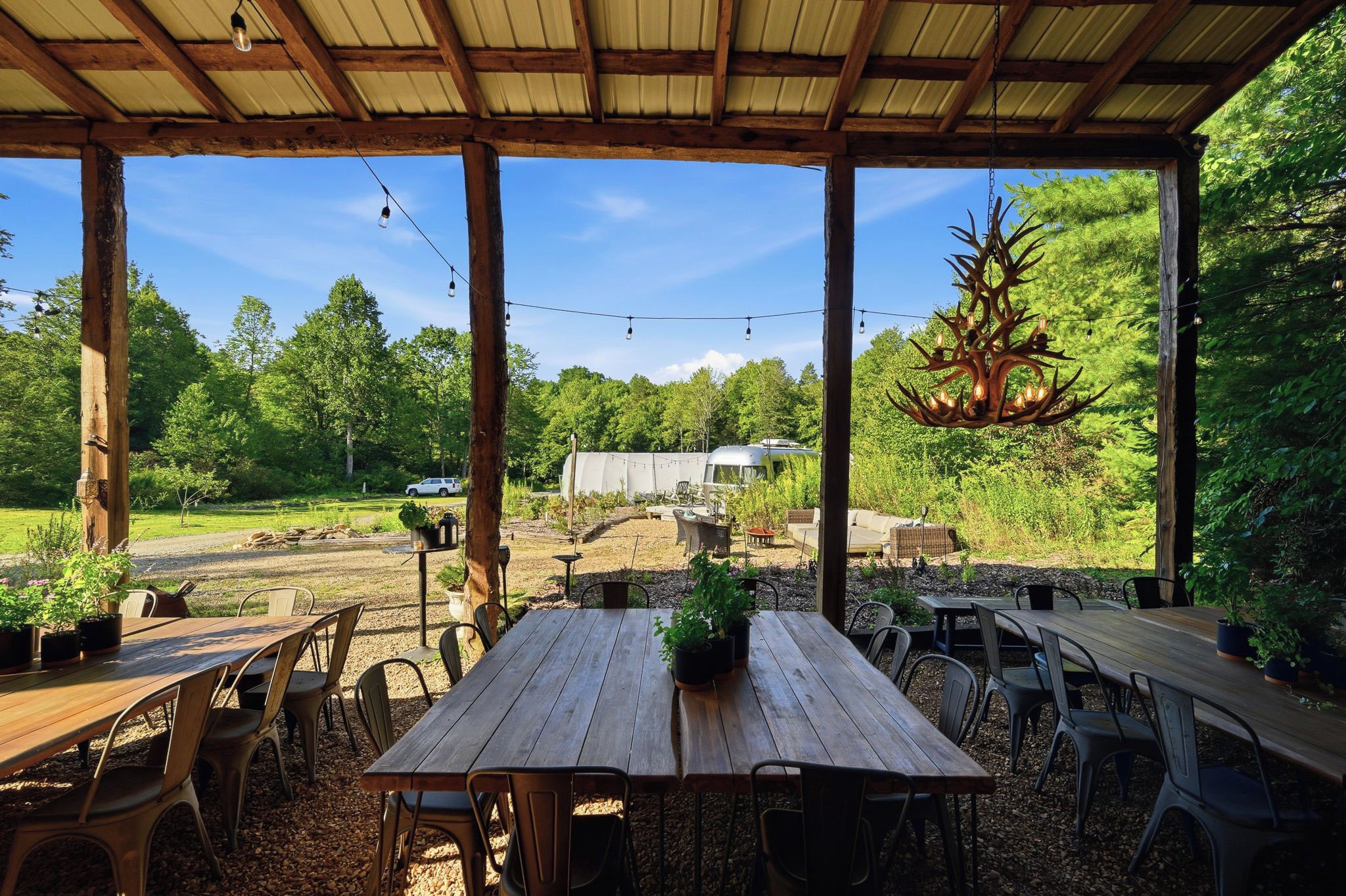 An outdoor patio with wooden tables and chairs, surrounded by greenery and a hanging chandelier.