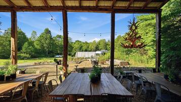 An outdoor patio with wooden tables and chairs, surrounded by greenery and a hanging chandelier.