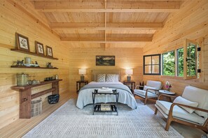 Ein Schlafzimmer einer Holzhütte mit einem Bett, Nachttischen, einem Sessel, einem kleinen Tisch und einem Fenster.