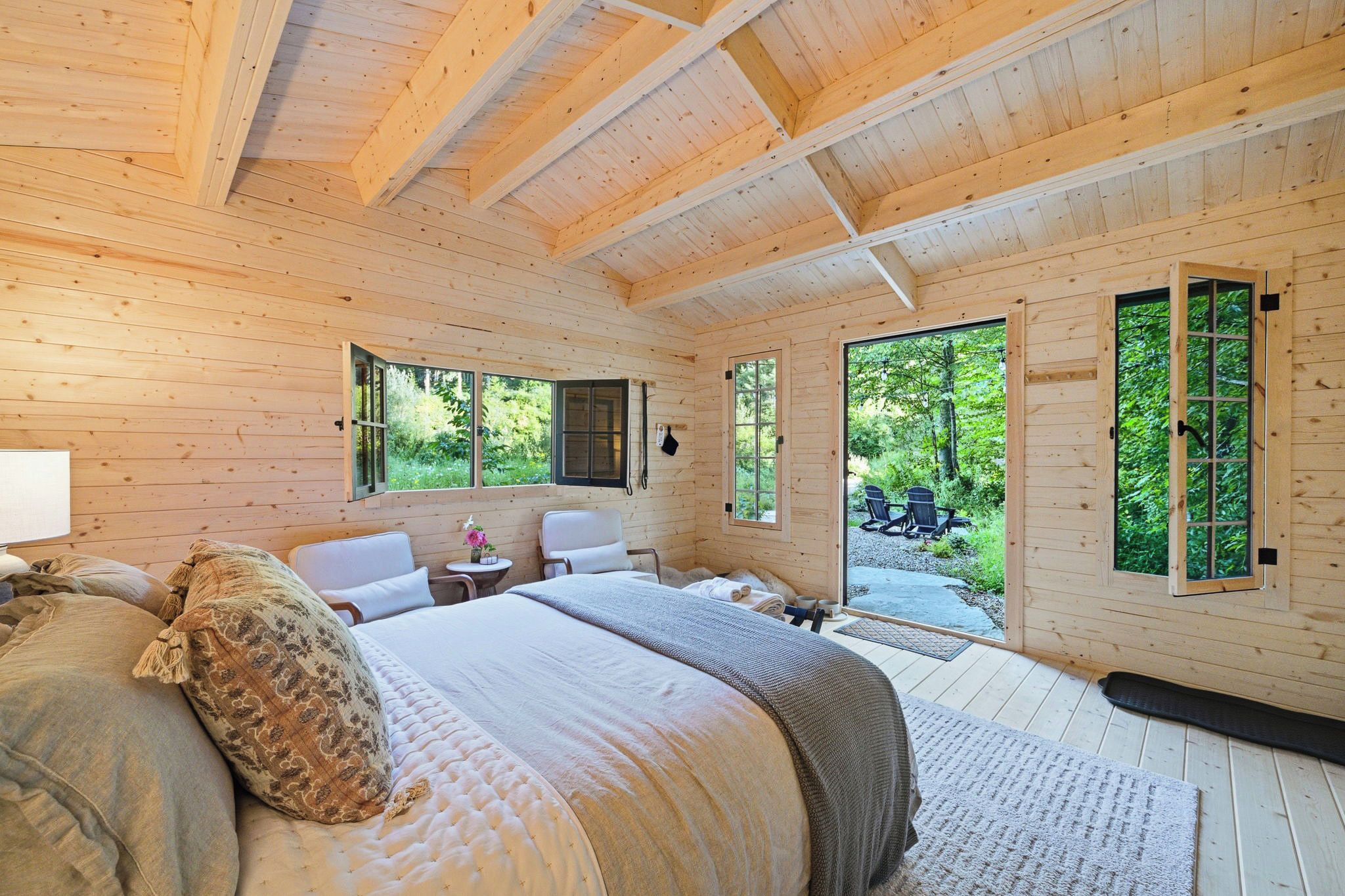 A wooden cabin bedroom with a bed, two chairs, a small table, and a window overlooking greenery.