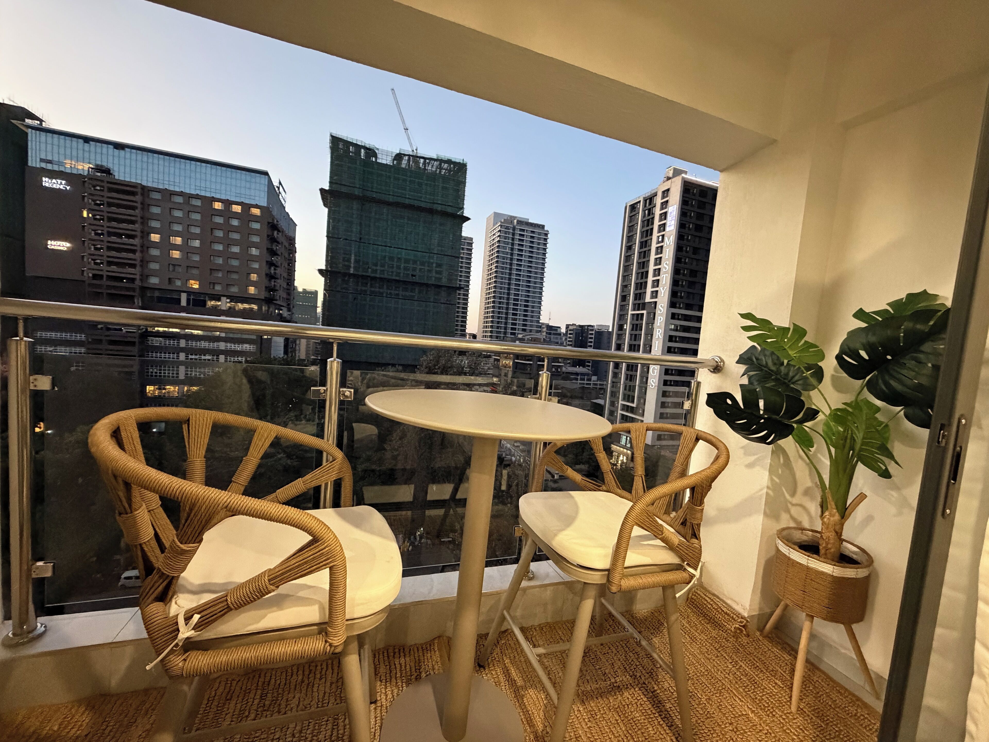 Un balcón con sillas de mimbre y una pequeña mesa redonda, con vistas a un paisaje urbano con rascacielos y una obra en construcción.