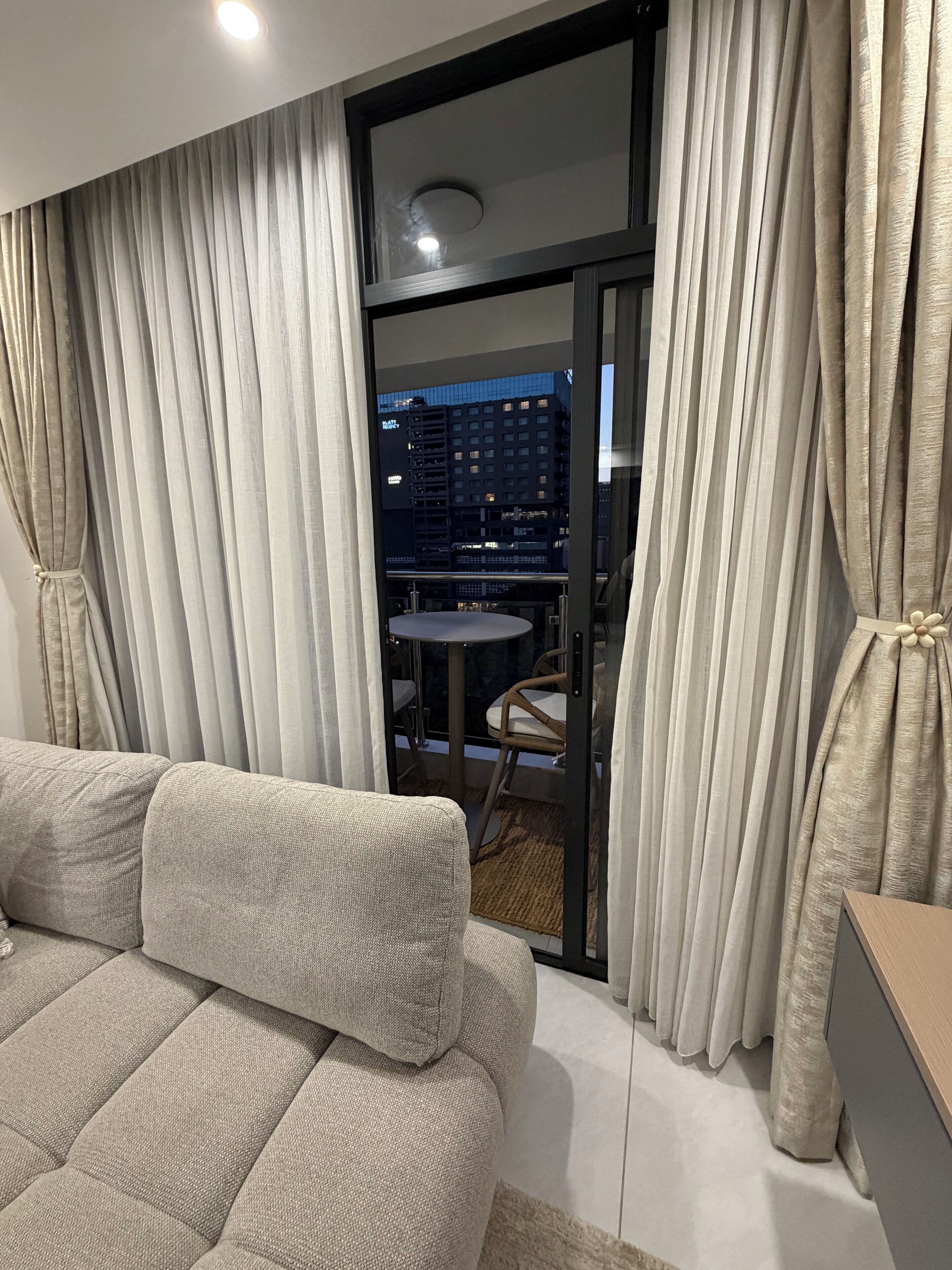 A modern living room with a sofa, curtains, and a view of a cityscape through a glass door.
