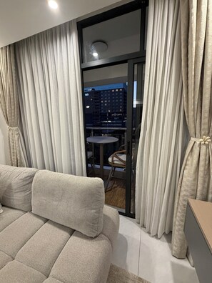 Ein modernes Wohnzimmer mit einer Couch, Vorhängen und Blick auf eine Stadtlandschaft durch eine Glasdoor.