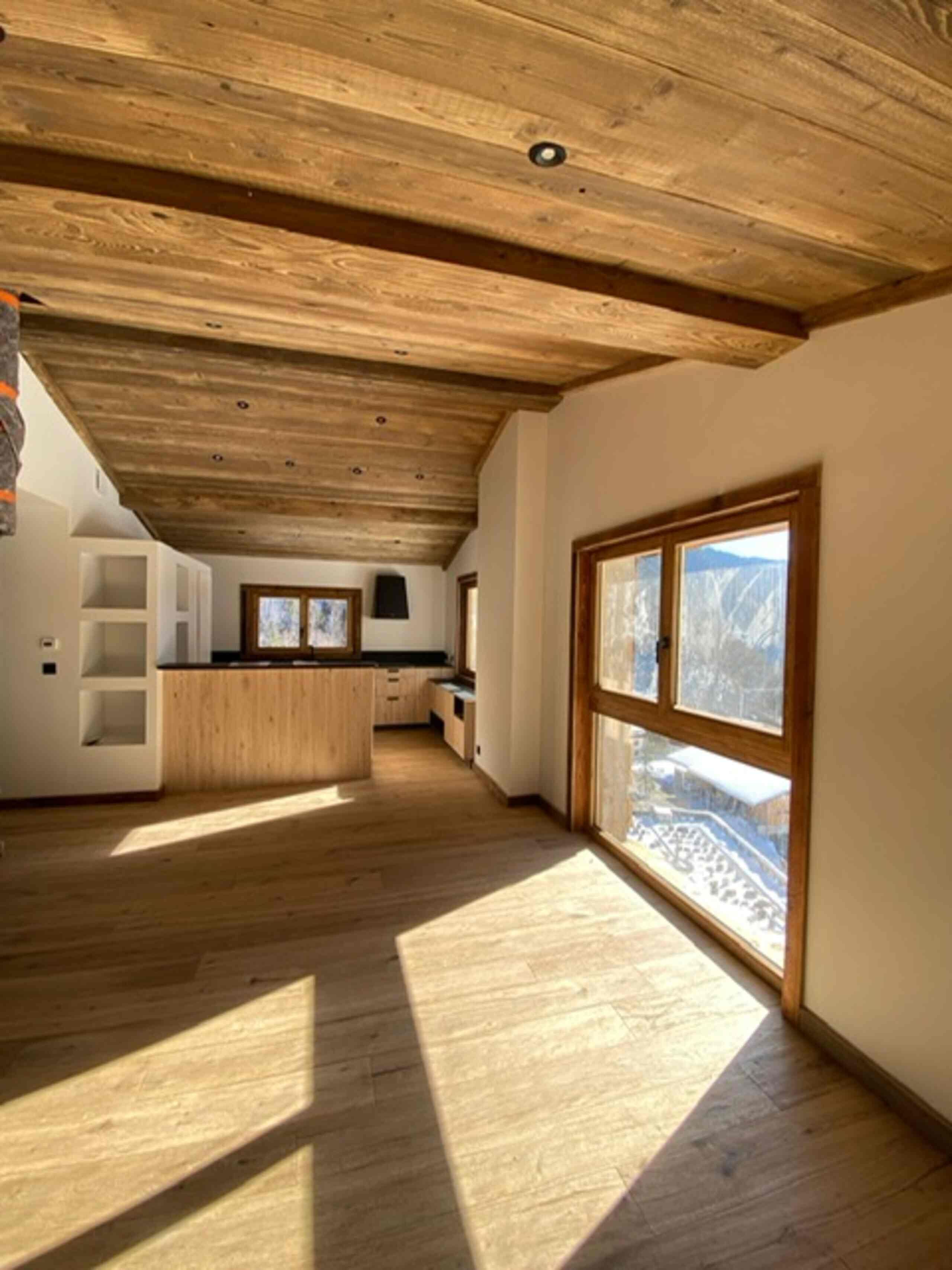 A spacious room with a wooden ceiling, a large window with a view, and a kitchen area with wooden cabinets.