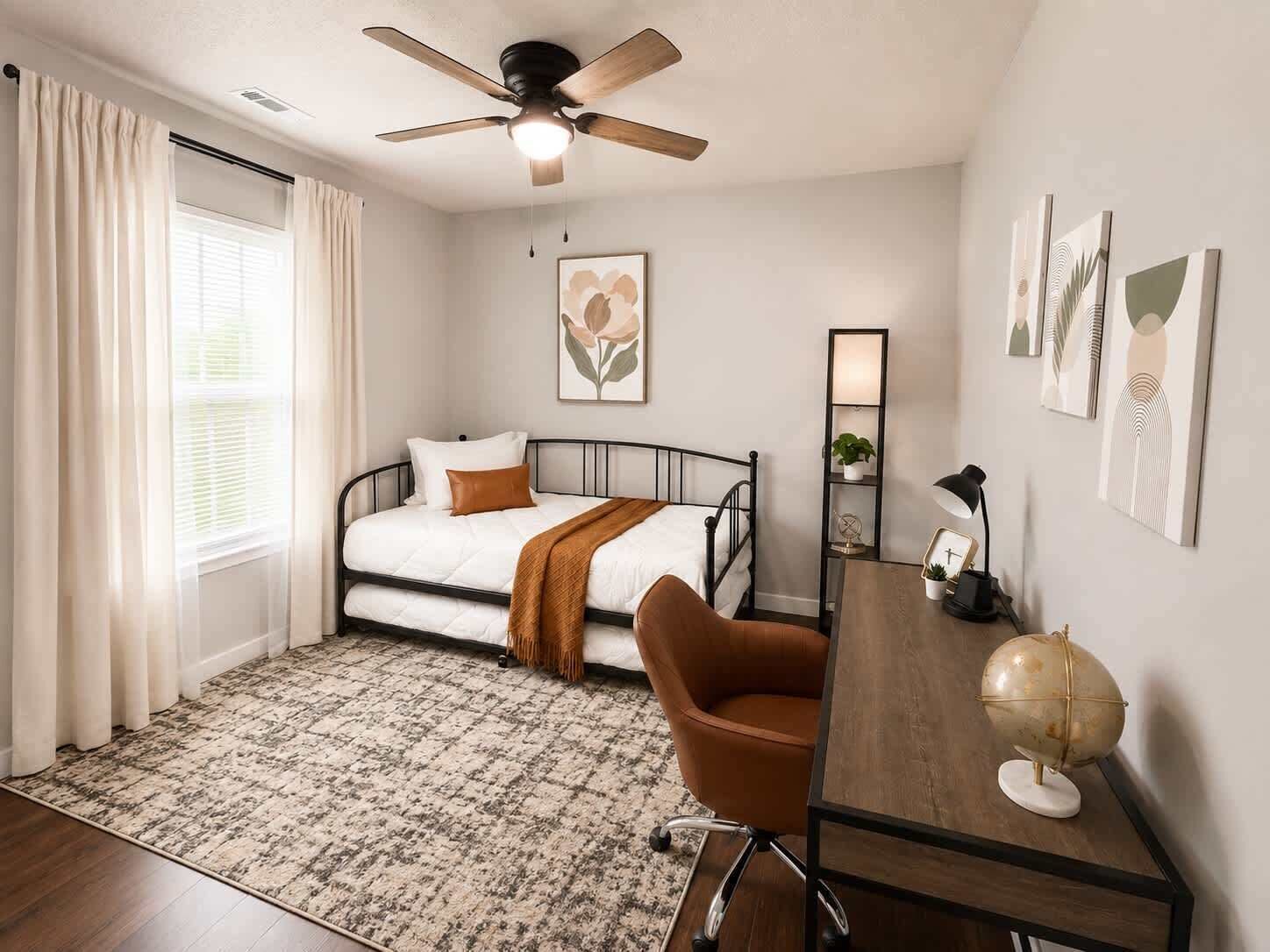 A bedroom with a bed, a desk, a chair, a ceiling fan, and artwork on the walls.