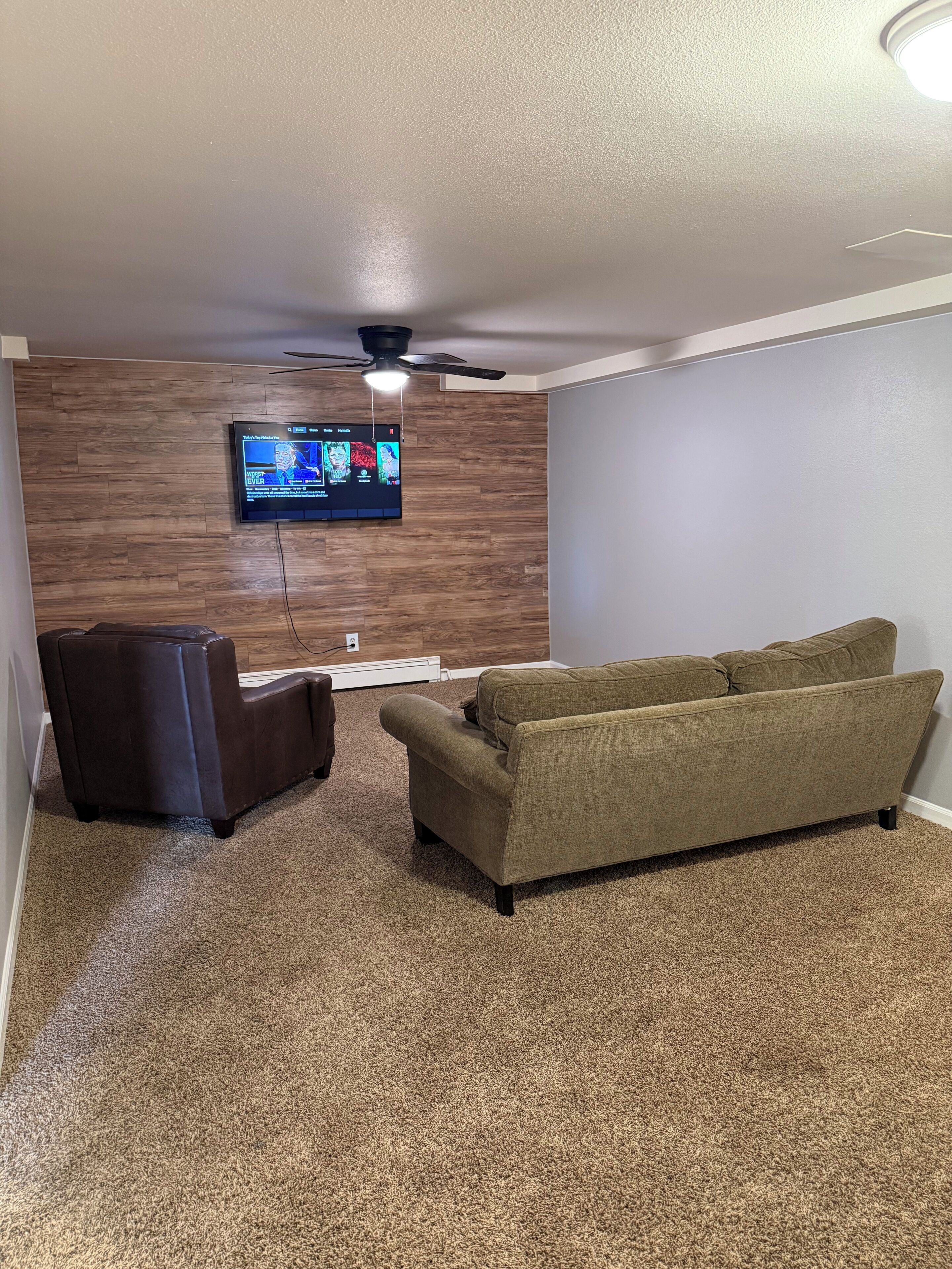 A living room with a flat-screen TV mounted on a wooden wall, a brown leather chair, a green sofa, and a ceiling fan.