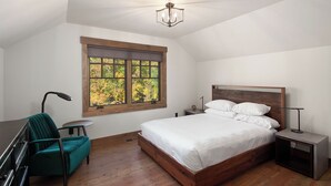 Ein Schlafzimmer mit einem hölzernen Bett, einem grünen Sessel, einem kleinen Tisch, einem Nachttisch und einem Fenster mit Blick auf Bäume.