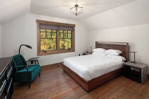 Un dormitorio con una cama de madera, una silla verde, una mesita pequeña, una mesita de noche y un ventanal con vistas a árboles.