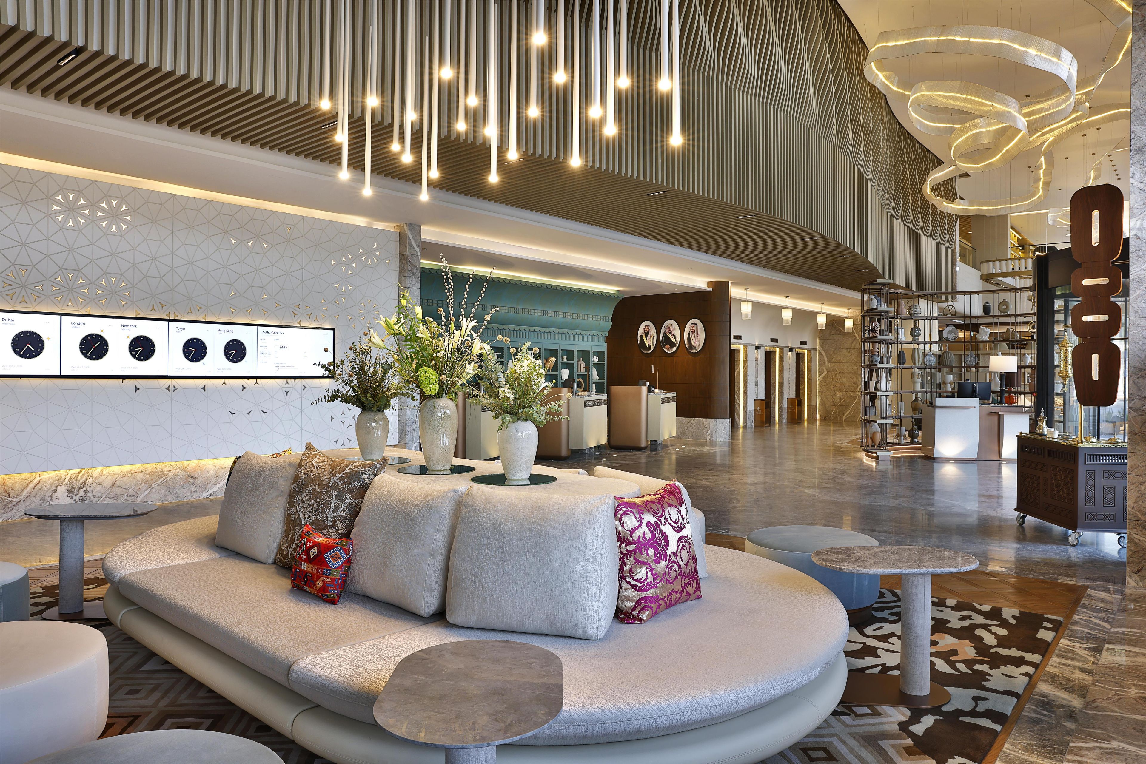 A modern hotel lobby with a seating area, a reception desk, and a decorative wall with clocks.
