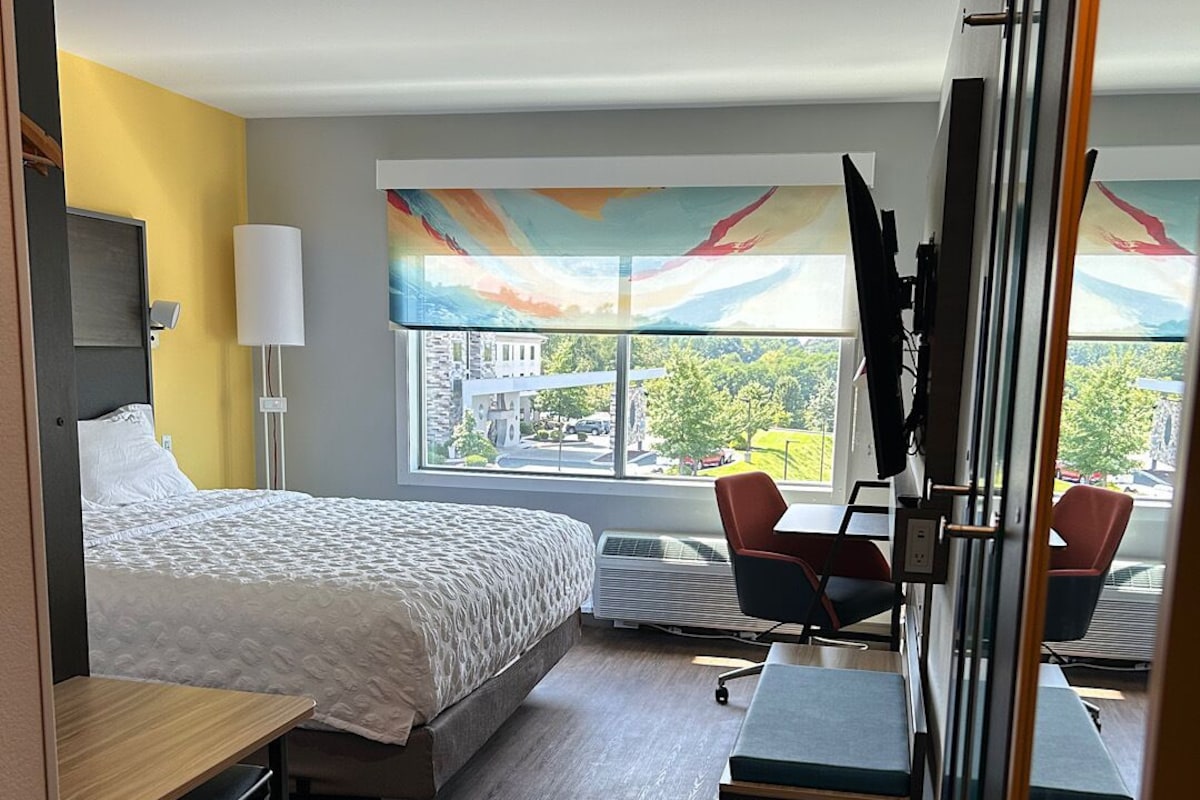 A hotel room with a bed, desk, chairs, and a large window with a view of buildings and trees.