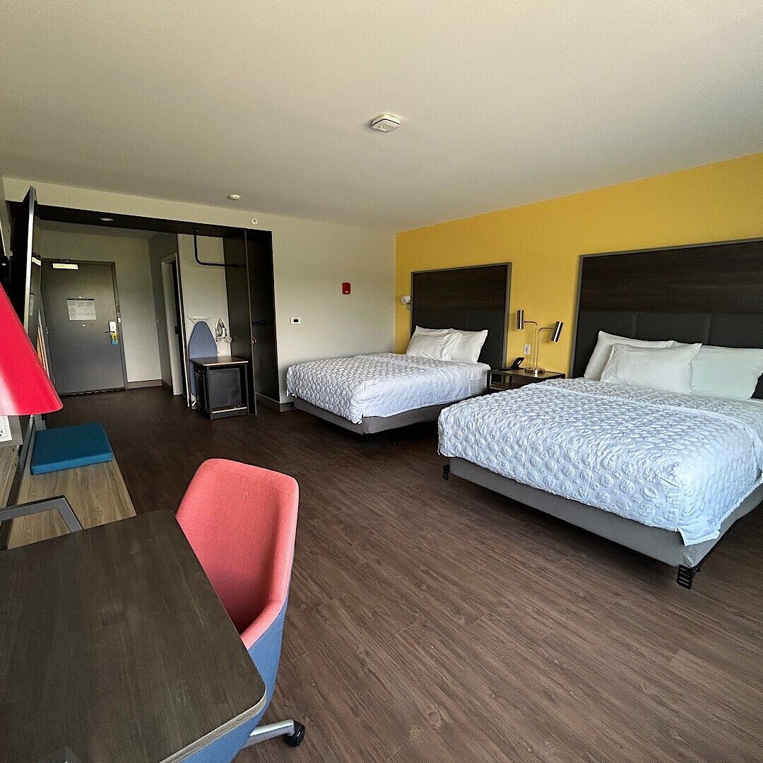A hotel room with two beds, a desk, a chair, and a door leading to a hallway.