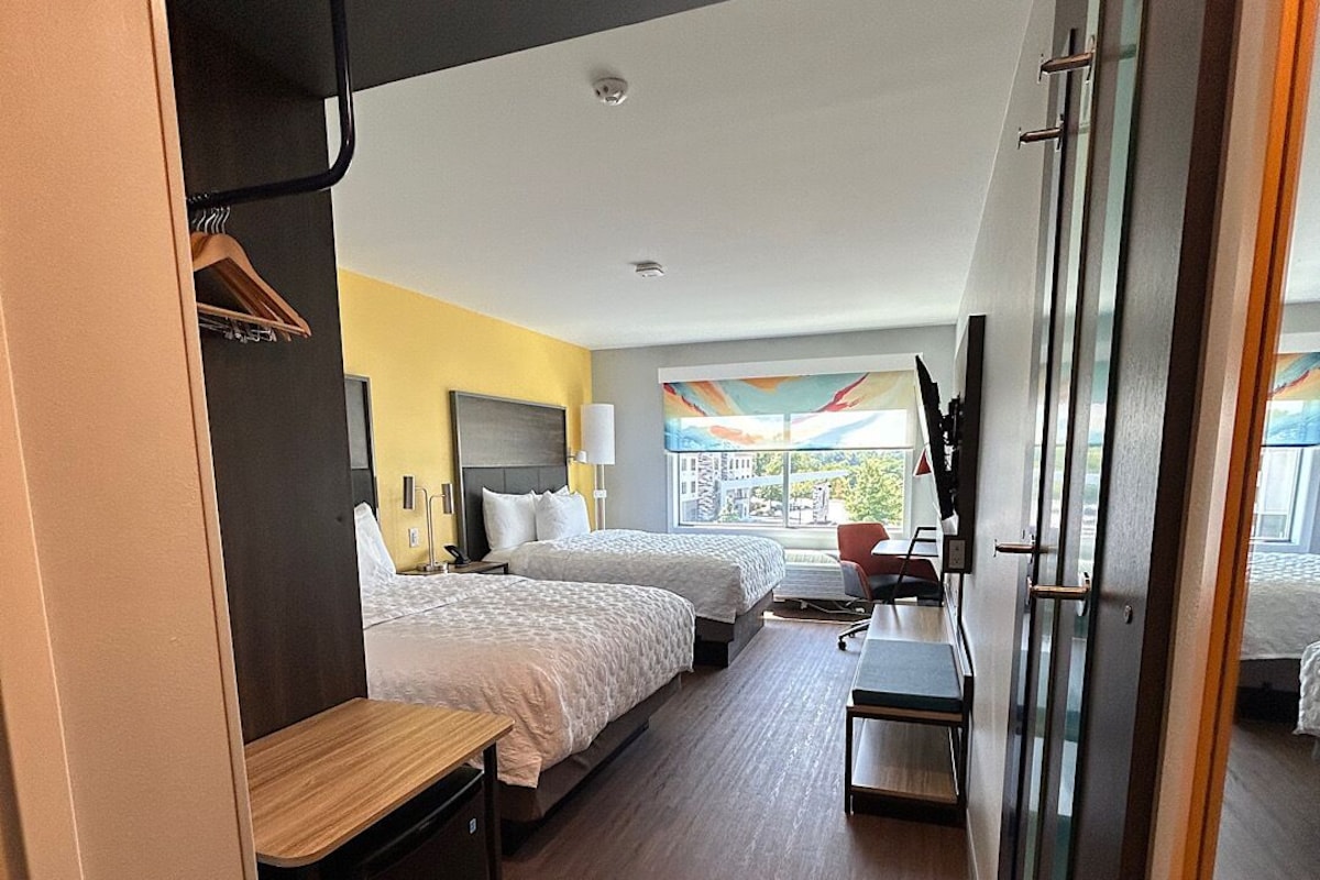 A hotel room with two beds, a desk, a chair, and a view of the outdoors.