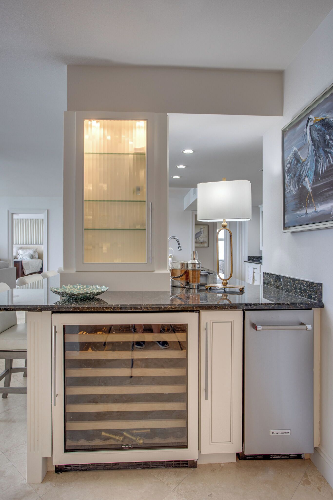 A modern kitchen with a built-in dishwasher, a large window with blinds, a countertop with a glass container, and a wall-mounted lamp.