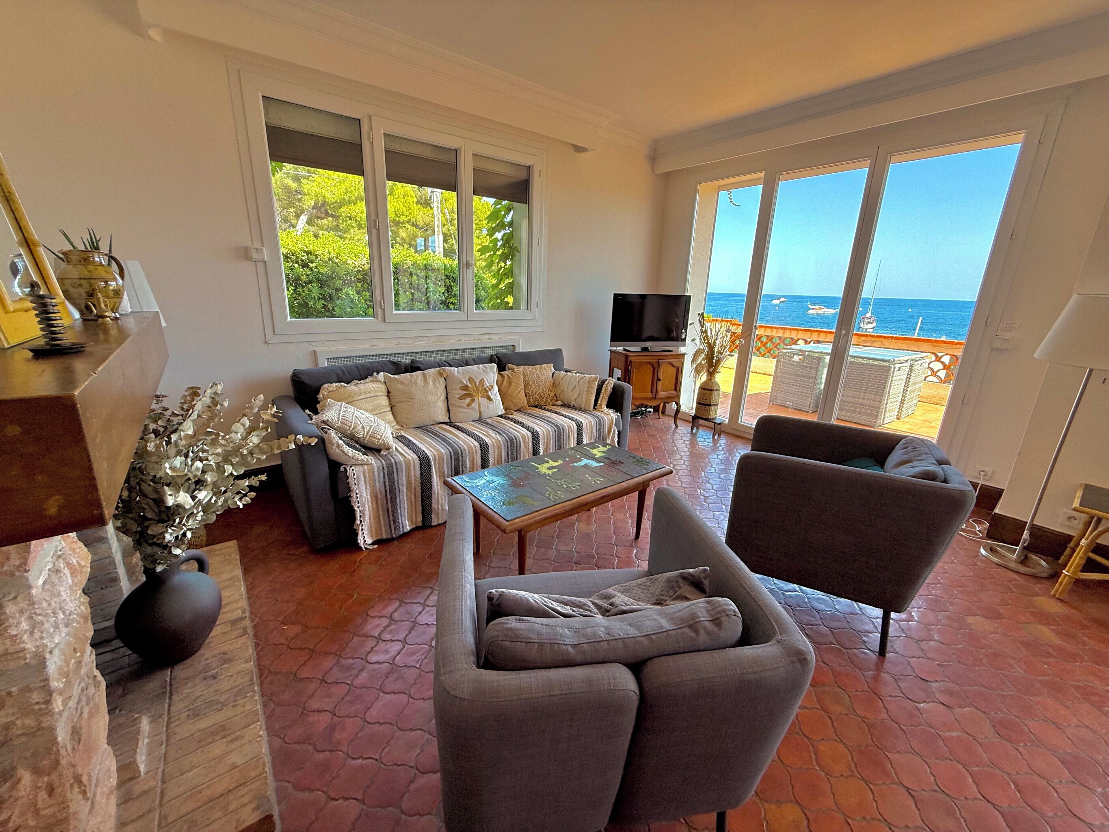 Un salon comprenant un canapé, des fauteuils, une table basse et un téléviseur. De grandes fenêtres offrent une vue sur la mer.