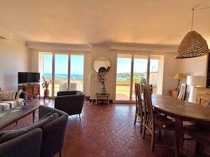 Ein geräumiger Wohnbereich mit Fernseher, Sitzgelegenheiten, einem Esstisch und Meerblick durch große Fenster.