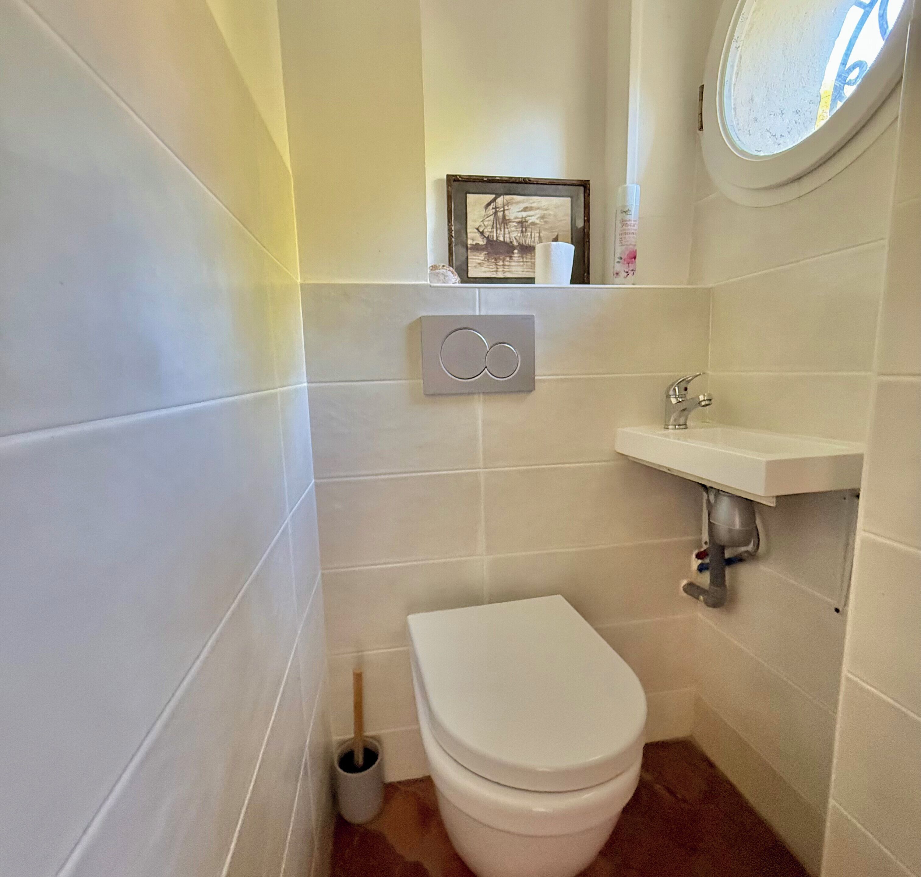 A small, clean bathroom with white tiles, a toilet, a sink, and a picture frame on the wall.