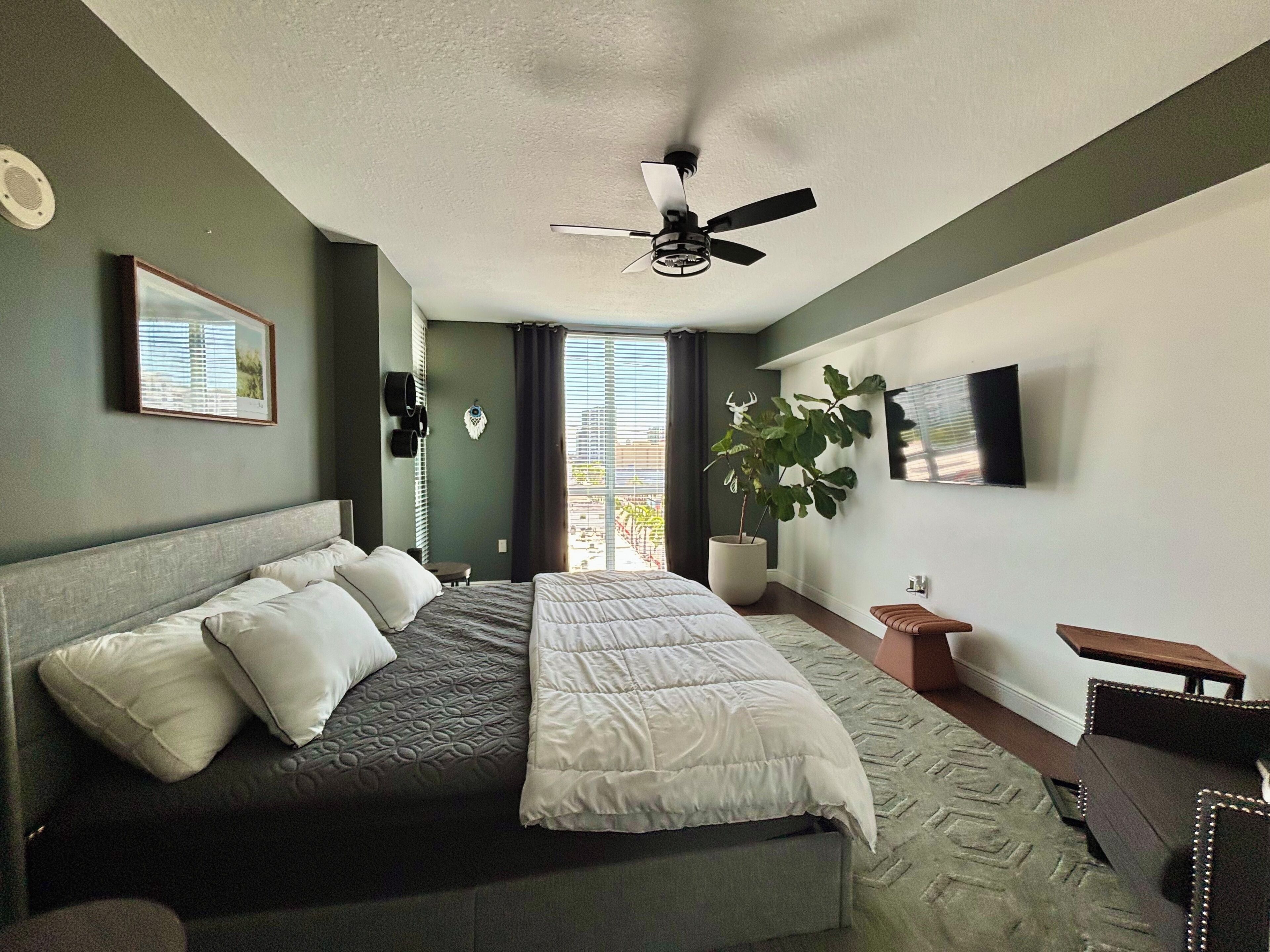 A modern bedroom with a large bed, a ceiling fan, a potted plant, and a view of the outdoors.